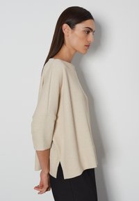 Beige gestrickter Pullover mit Dreiviertelärmeln, Bootsausschnitt, seitlichen Schlitzen und lockerer Passform. Strukturierter Stoff mit dezentem Glanz.