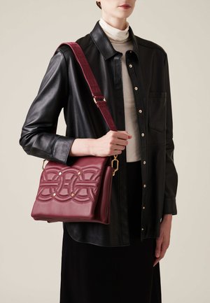 Burgunderfarbene Ledertasche mit geprägtem Muster, goldenen Hardware-Details und verstellbarem Tragegurt, gehalten gegen ein dunkles Outfit. Glatte Textur.