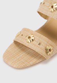 Beige woven sandals with two straps, adorned with gold flower embellishments and small gold studs. Square toe with textured surface.
