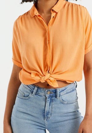 Person wearing a short-sleeve orange button-up shirt tied at the waist and high-waisted light blue jeans against a white background.