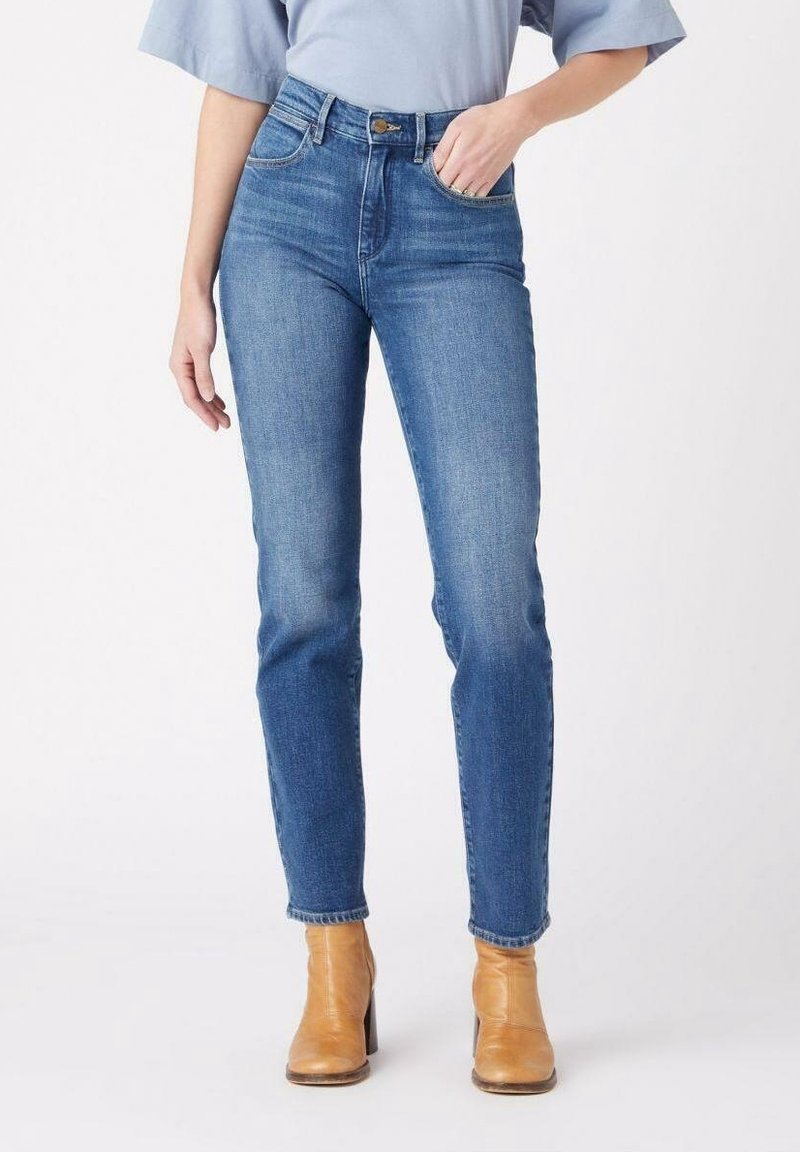 Jeans de tiro alto en azul, fabricados en denim, con una silueta ajustada, diseño de cinco bolsillos y un ligero desvanecimiento. Combinados con botas de tobillo marrones.