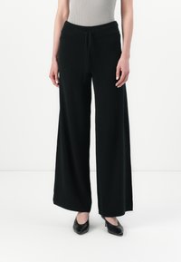 Pantaloni neri a gamba larga realizzati in tessuto morbido, con vita regolabile e vestibilità rilassata, abbinati a tacchi neri.