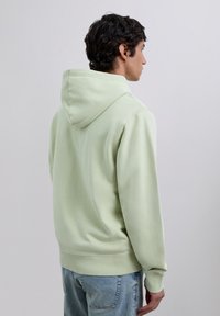 Man facing sideways wears light green hooded sweatshirt and light blue jeans against plain light gray background.