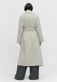 Lång, ljusgrå ullrock med bälte i midjan, vida ärmar och ryggslits. Tillsammans med lösa, mörkgrå byxor. Minimalistisk design.