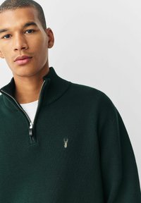 Suéter verde con cremallera y cuello acanalado, tela texturizada, y un pequeño logo bordado en el pecho. Camiseta blanca se ve debajo.