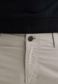 Pantalones beige con un botón metálico que muestra un logo, combinados con una prenda de tela acanalada negra por encima de la cintura.
