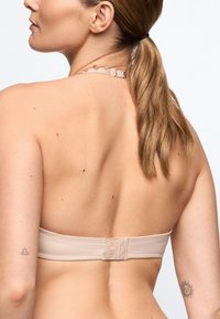Femme aux cheveux châtain clair en queue de cheval, portant un soutien-gorge bandeau couleur peau, un collier de coquillage, et des tatouages sur les deux bras supérieurs, vue de dos.