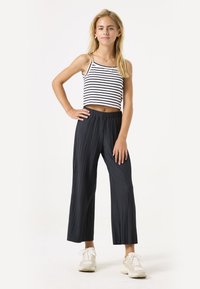 Zwarte geplisseerde culottes met een elastische tailleband, gecombineerd met een wit en zwart gestreept crop top. Model staat met één hand op de heup.
