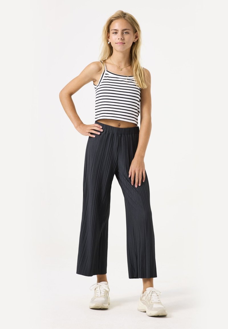Zwarte geplisseerde culottes met een elastische tailleband, gecombineerd met een wit en zwart gestreept crop top. Model staat met één hand op de heup.