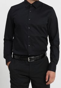 Svart långärmad skjorta med knappkrage, med en mjuk textur och subtil glans, kombinerad med svart bälte och byxor.