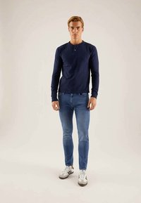 Tengerészkék, hosszú ujjú henley póló világoskék skinny farmernadrággal és szürke sportcipővel párosítva, kerek nyakkal és finoman texturált anyaggal.