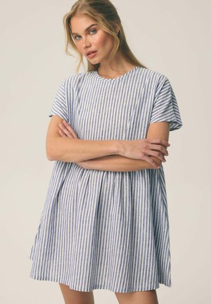 Młoda kobieta o blond włosach, ubrana w krótką sukienkę w niebiesko-białe pionowe paski, z założonymi ramionami, na jednolitym tle.