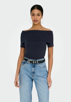 Femme aux cheveux foncés portant un haut bleu marine à épaules dénudées, un jean taille haute bleu clair, une ceinture noire et de grandes boucles d'oreilles en anneau, faisant face à l'avant.