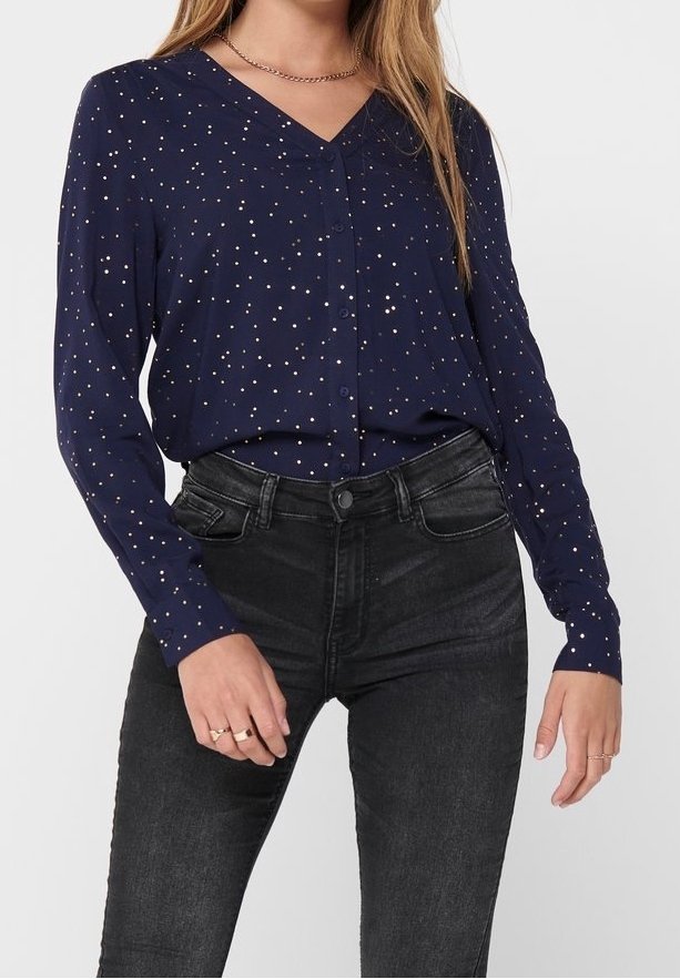 Femme portant un chemisier bleu marine à manches longues, boutonné, à pois blancs, et un jean noir taille haute, debout, mains détendues.
