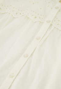 Weißes Kleid mit besticktem Spitzenbesatz, ausgestattet mit Knopfverschluss und weichem, transparentem Stoff, der von zarten Blumenmustern und gewellten Kanten akzentuiert wird.