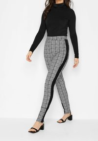 Svart polotröja i kombination med grå rutiga leggings som har en svart sidopanel. Leggingsen är åtsittande med en stretchig textur, och modellen bär svarta sandaler.