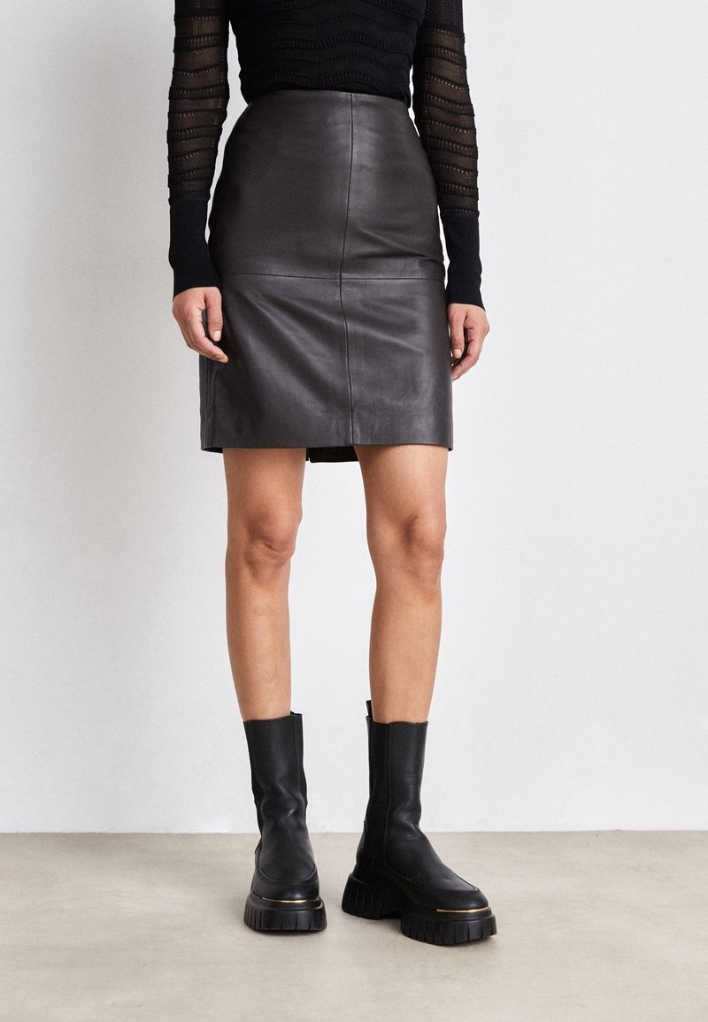 Sort lædernederdel med høj talje og tætsiddende design, med vandrette syninger, kombineret med chunky sorte støvler med hæl. Glat tekstur.