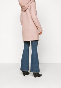 Cappotto con cappuccio rosa chiaro a taglio dritto; tessuto testurizzato, di lunghezza media, abbinato a jeans a campana blu e stivali neri con tacco.