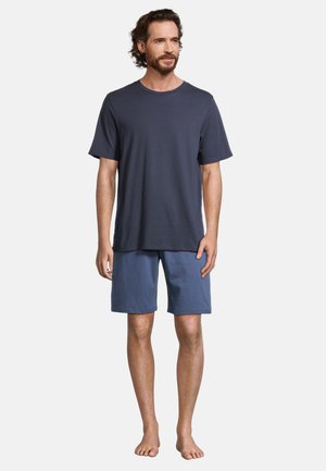 Mężczyzna z ciemnymi włosami i brodą, ubrany w granatowy t-shirt z krótkim rękawem i niebieskie spodenki, stoi boso na jednolitym tle.