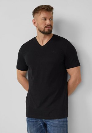 Mann mit Bart, der ein schwarzes V-Ausschnitt-T-Shirt und blaue Jeans trägt, steht vor einer schlichten hellgrauen Wand und schaut nach links.