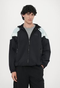 Črna zip-up jakna z svetlo modrimi ramenskimi poudarki in teksturiranimi črtami. Ima kapuco, dvojna sprednja žepa in elastične manšete.