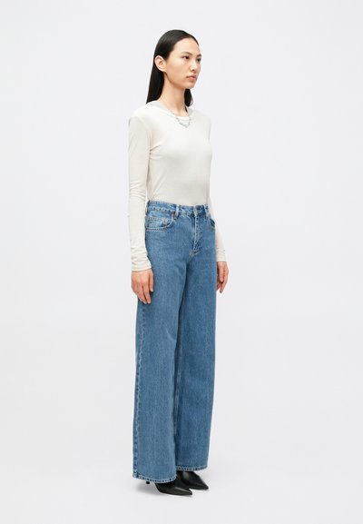 Ανοιχτόχρωμο τοπ με μακριά μανίκια συνδυασμένο με ψηλόμεσο, φαρδύ μπλε τζιν και μαύρα μυτερά τακούνια. Απλός σχεδιασμός με ελάχιστες λεπτομέρειες.