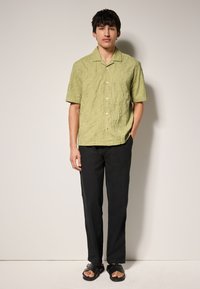 Lichtgroen T-shirt met korte mouwen en een gestructureerd patroon, met knopen aan de voorkant. Zwarte broek, platte zwarte sandalen met bandjes, staand op een lichte ondergrond.