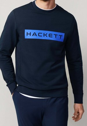 Man draagt een marineblauwe trui met een blauwe rechthoekige logoband over de borst met de tekst "HACKETT."
