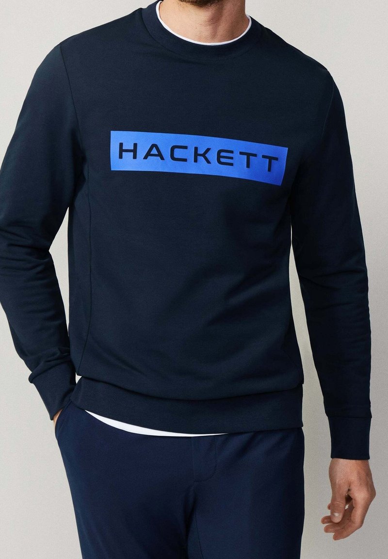Homme portant un sweat-shirt bleu marine avec une barre de logo rectangulaire bleue sur la poitrine affichant le mot "HACKETT."