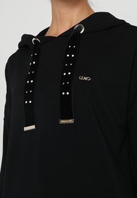 Sudadera negra con cordón, con herrajes dorados brillantes y acentos de terciopelo. El logo está sutilmente colocado en la parte delantera. Textura suave.