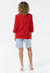 Blazer en lin rouge à manches trois-quarts, porté avec un short en jean bleu clair, associé à des sandales à talons haut beiges. Texture lisse.