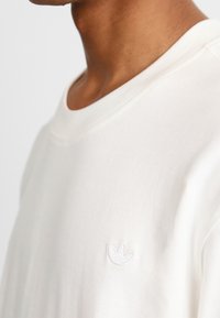 White cotton sweatshirt with a crew neck, featuring a small white embroidered logo on the chest and a smooth, textured finish.