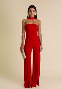 Rode strapless jumpsuit met een aansluitende bovenkant, flare benen, zijsplitten en een zachte textuur. Gecombineerd met een metallic clutch en statement oorbellen.
