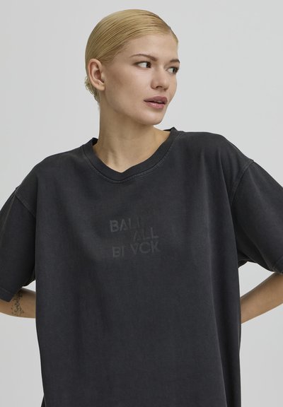 Szőkére festett hajú nő, haja hátrafésülve, bő fekete pólót visel halvány felirattal, jobbra néz egyszínű háttér előtt.