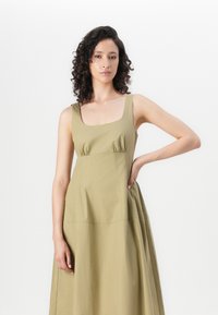 Brezrokavni olivno zeleni plašč z kvadratnim izrezom, nagubanimi bokovi in takočnim krilom, ki se ponaša z gladkim materialom in subtilnimi šivi.