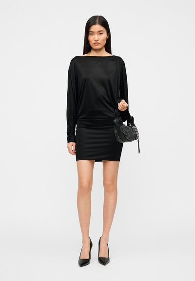 Μαύρο μακρυμάνικο τοπ σε συνδυασμό με στενή μαύρη μίνι φούστα. Το μοντέλο κρατά μια μαύρη τσάντα με καρφιά και φοράει μυτερά μαύρα τακούνια.