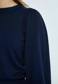 Námorně modrý pletený svetr s kulatým výstřihem a nadýchanými dlouhými rukávy, s hladkou texturou a žebrovaným lemem. Jednoduchý design bez vzorů.