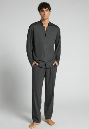 Jeune homme debout pieds nus, portant une chemise boutonnée à manches longues gris foncé et un pantalon assorti sur un fond clair uni.