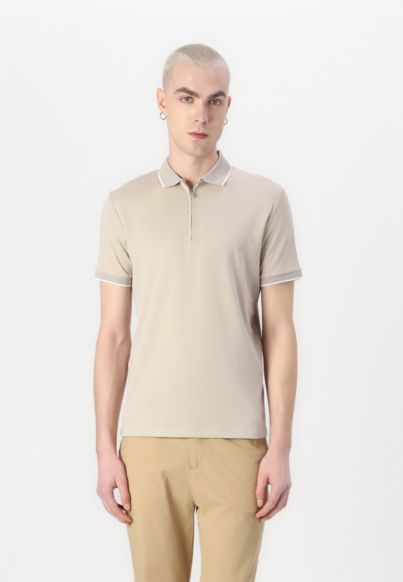 Beige polo skjorte med glidelås krage, kontrasterende hvit kanting på kragen og ermene, korte ermer og en glatt tekstur.