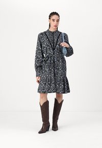 Abito floreale nero con maniche lunghe, vita cintrata e orlo sfrangiato. Abbinato a stivali da cowboy marroni. Una piccola borsa blu è indossata sulla spalla.