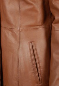 Bruine leren jas met een soepele textuur, een slank ontwerp en een zijzak met een smalle opening. Verticale stiknaaddetails zichtbaar.