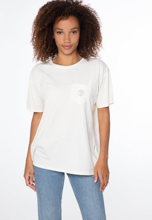 Cremefarbene Baumwoll-T-Shirt mit kurzen Ärmeln, entspanntem Schnitt und einer linken Brusttasche mit einem runden Logo-Print. Kombiniert mit blauen Jeans.
