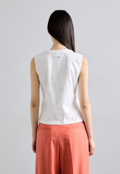 White sleeveless top made of cotton, featuring a round neckline and a small red logo detail at the back, paired with coral trousers.