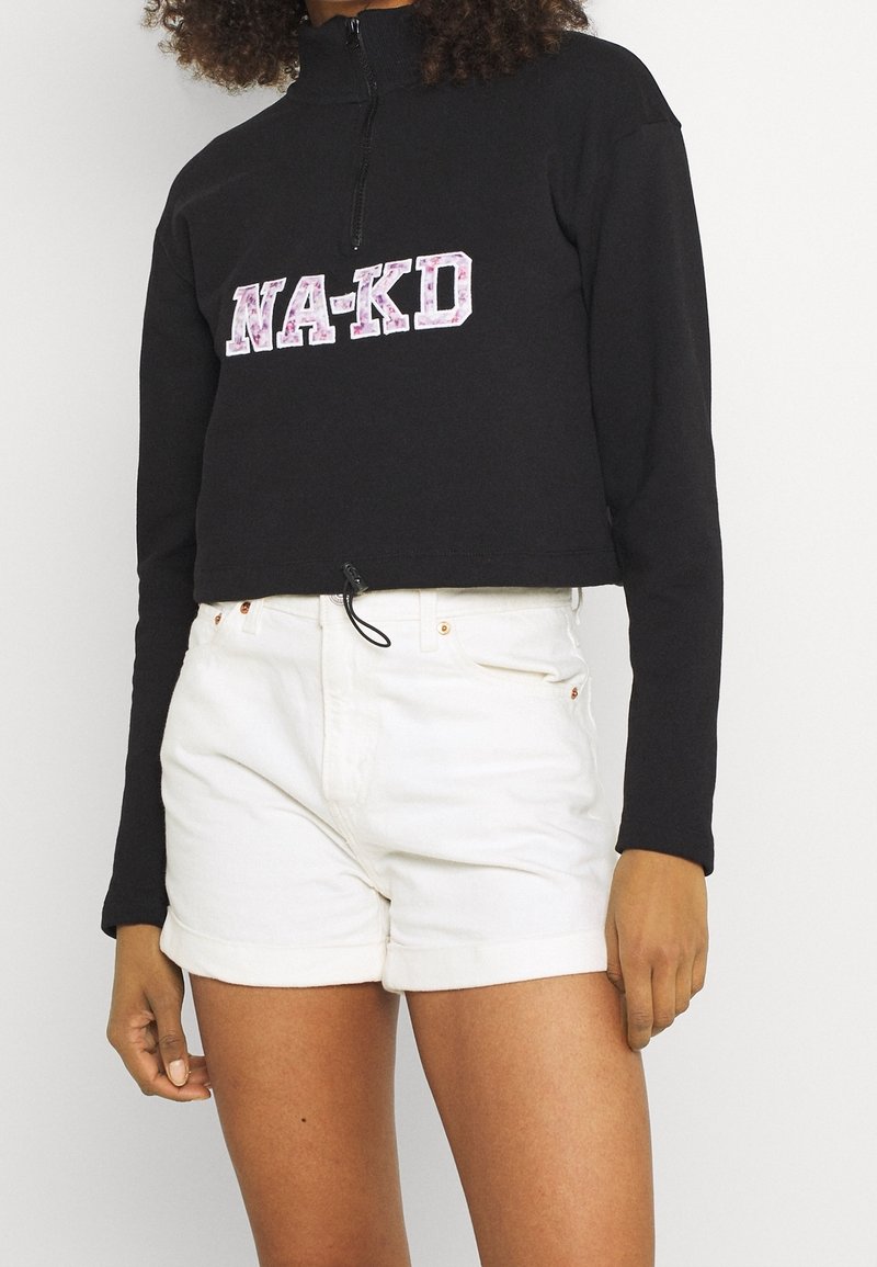 Felpa nera corta con colletto alto, con un logo "NA-KD" in rosa e bianco, abbinata a pantaloni corti bianchi. Tessuto testurizzato con cordino.