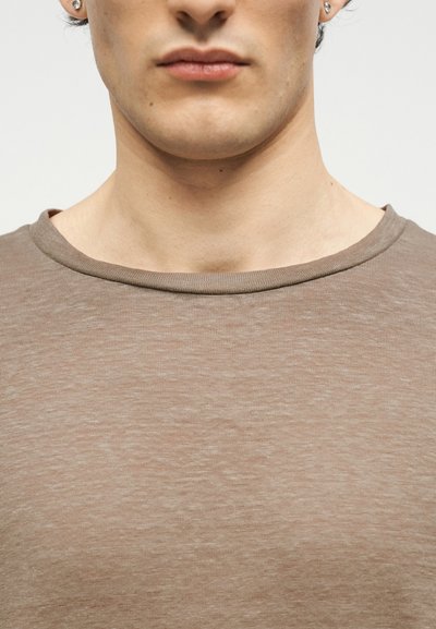 Κοντινή λήψη του κάτω μέρους του προσώπου και του λαιμού ενός ατόμου που φορά σκουλαρίκια από διαμάντια και ένα ανοιχτό καφέ μπλουζάκι με στρογγυλή λαιμόκοψη, με ουδέτερο φόντο.