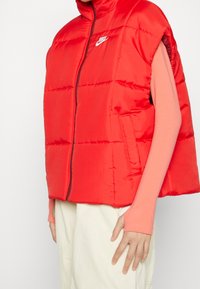 Röd vadderad väst med ärmlös design, hög krage och sidofickor. Har en framficka med dragkedja och en liten vit logotyp p å bröstet.