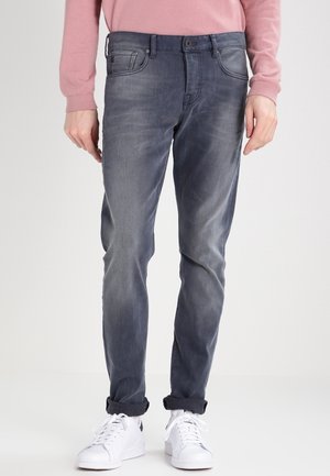 Personne portant un jean slim gris délavé, un haut à manches longues rose et des revers retroussés avec des baskets blanches sur un fond uni.
