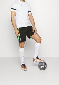 Biała koszulka Nike z czarnymi paskami, czarne szorty z zielonymi akcentami i logo Liverpoolu, białe skarpetki, pomarańczowo-granatowe buty piłkarskie, czarna piłka.