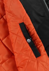 Kurtka pikowana z intensywnie pomarańczowym wnętrzem i czarnym zewnętrzem, o eleganckim designie i srebrnym zapięciem na kieszeni.