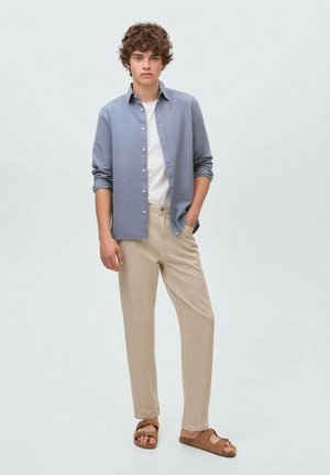 Chemise bleu clair à manches longues, t-shirt blanc, pantalon beige et sandales marron. La chemise a une coupe décontractée et des manches retroussées.
